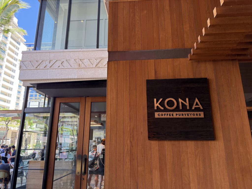 The storefront entrance of Kona Coffee Purveyors on a sunny day. It features a warm, wood-paneled wall with the shop's dark wood sign.