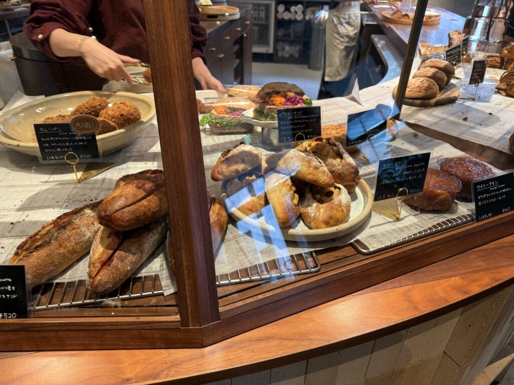 A variety of breads, including curry bread, sweet potato baguettes, and kouign-amann, are displayed in a wooden bakery showcase, with staff working in the background.