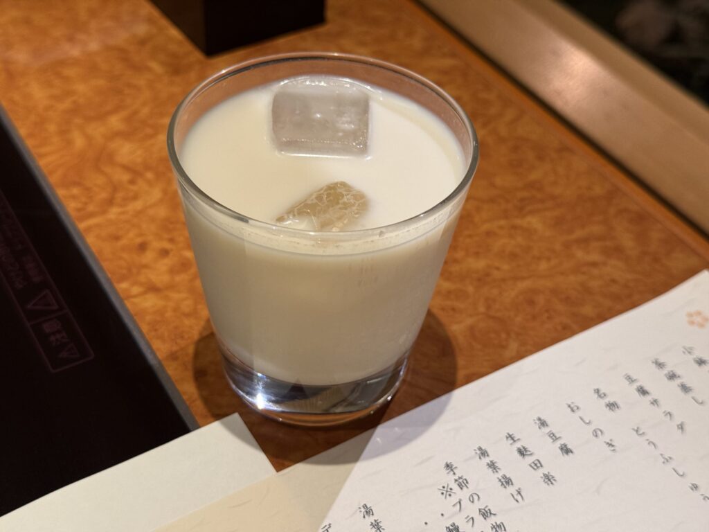 A glass of cold soy milk amazake with ice on a wooden table, next to a Japanese menu.