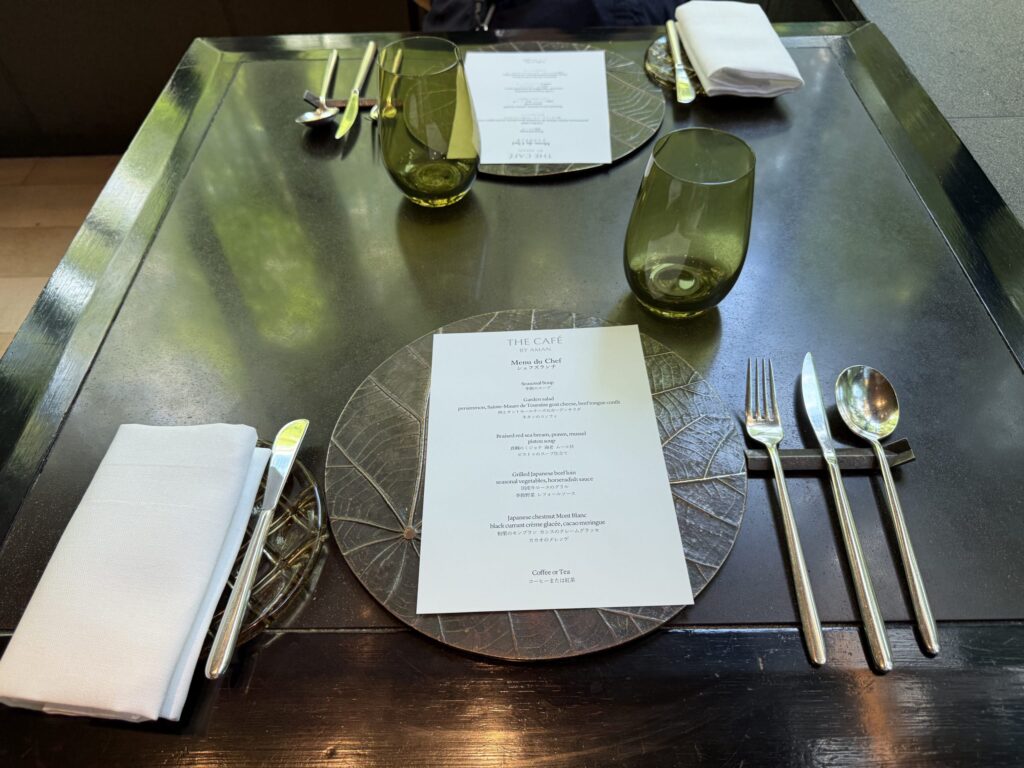 An elegant table setting for two at The Café by Aman, with a menu for the chef's lunch placed on a dark, leaf-patterned plate, alongside a green glass and silverware on a black table reflecting greenery.