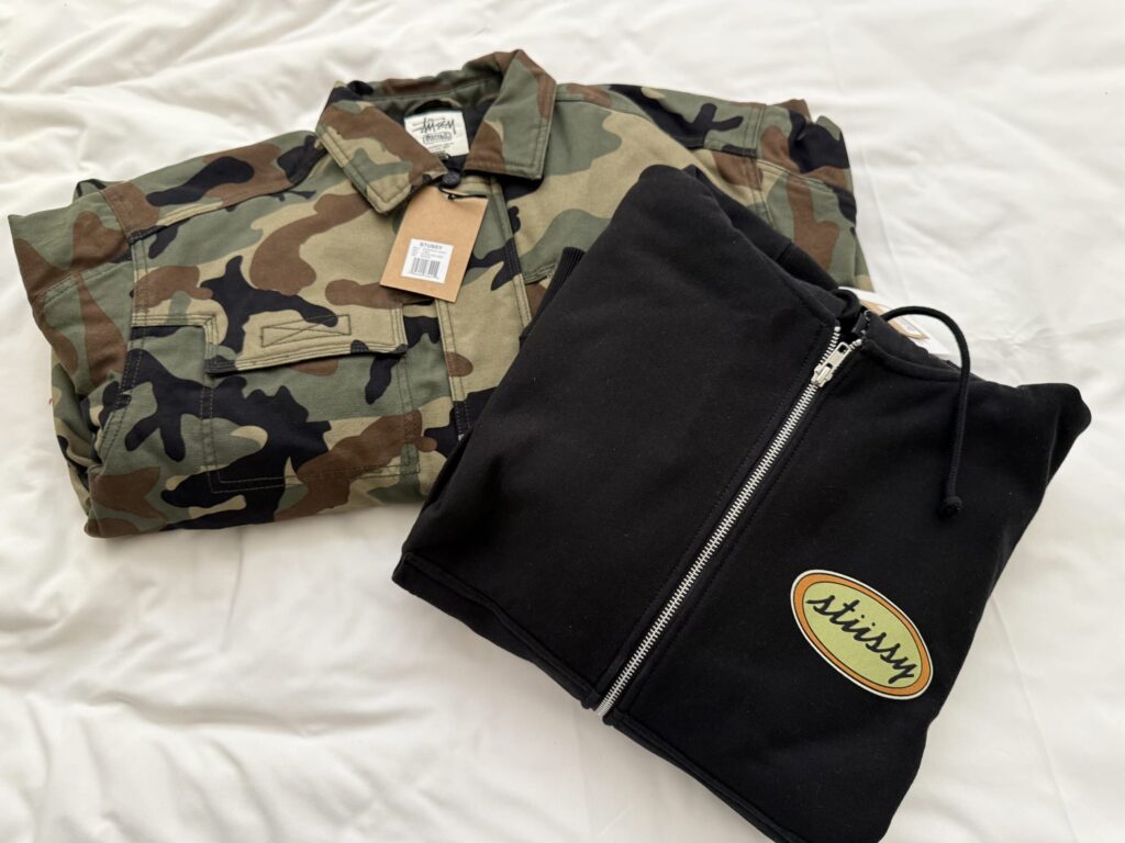 白い布の上に置かれたStussyの迷彩柄ジャケットと、胸にロゴワッペンが付いた黒いジップパーカー。