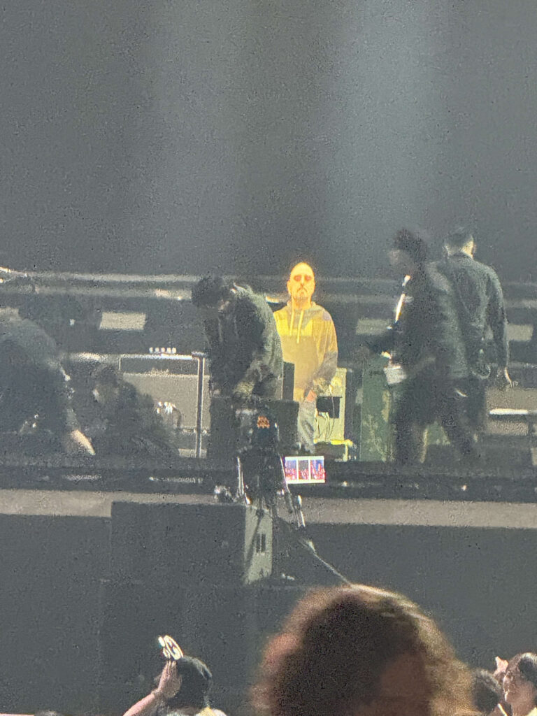 In the center of a dimly lit concert stage, a life-sized cardboard cutout of a man is illuminated by a yellow spotlight. Silhouettes of other band members are around it, and the heads of the audience are visible in the foreground.