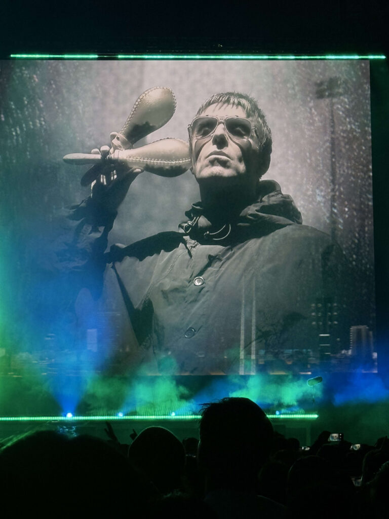 A black-and-white photo of Liam Gallagher in a parka and sunglasses holding maracas is displayed on a giant screen on a concert stage, with the silhouettes of the audience in the foreground.