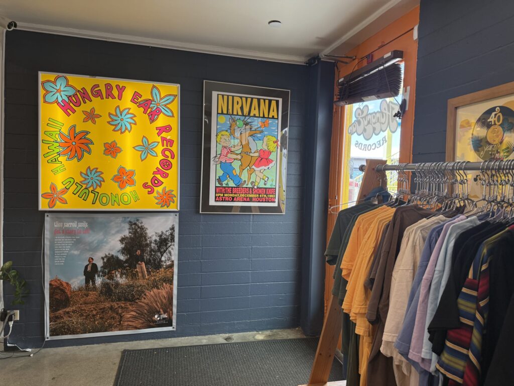 The entrance area of a record store. On the wall are a yellow lightbox poster for 'HUNGRY EAR RECORDS' and a Nirvana concert poster, with a clothing rack of various colored T-shirts placed next to them.