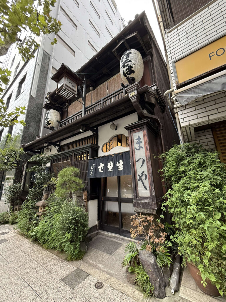 The charming wooden exterior of the long-established soba restaurant 'Kanda Matsuya' in Kanda, Tokyo. White paper lanterns and a black noren curtain hang at the entrance, surrounded by lush green plants.
