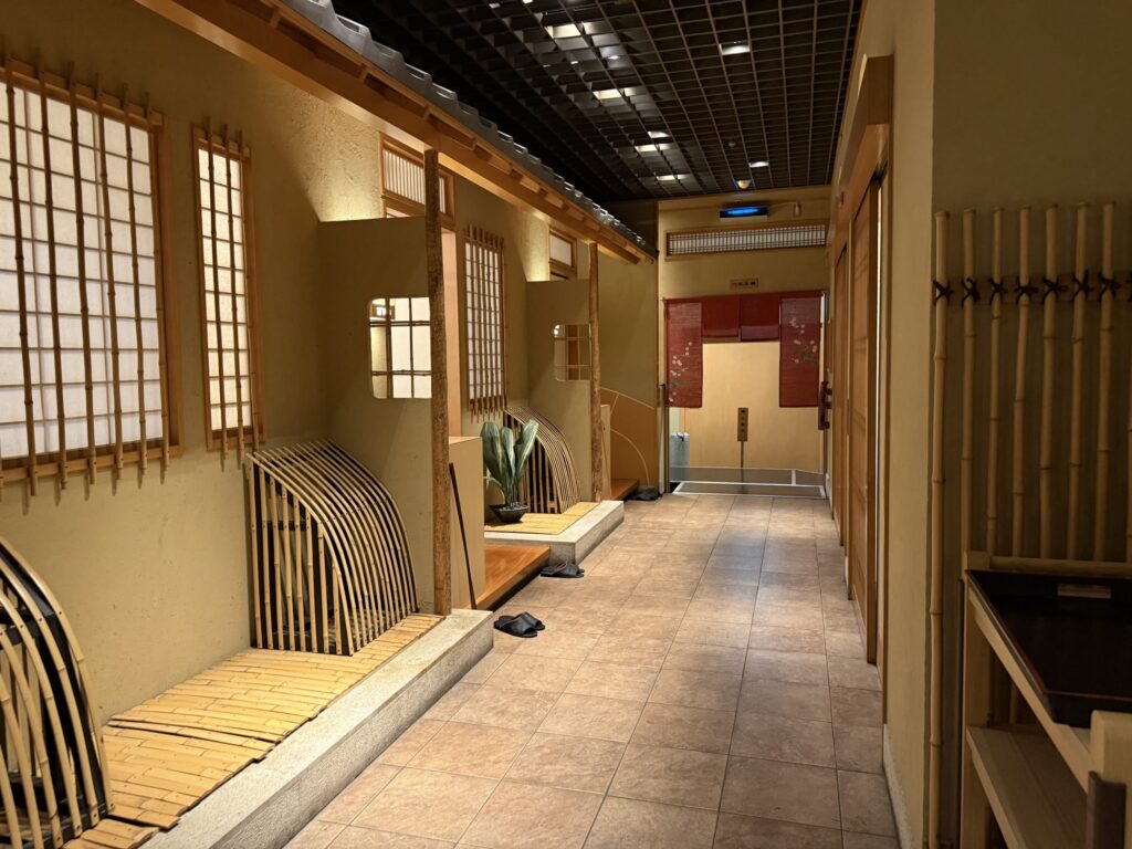 A hallway in a Japanese-style restaurant illuminated by warm lighting. Along the earthen-style walls are shoji windows with bamboo lattices and curved bamboo benches. A red noren curtain hangs in the distance.