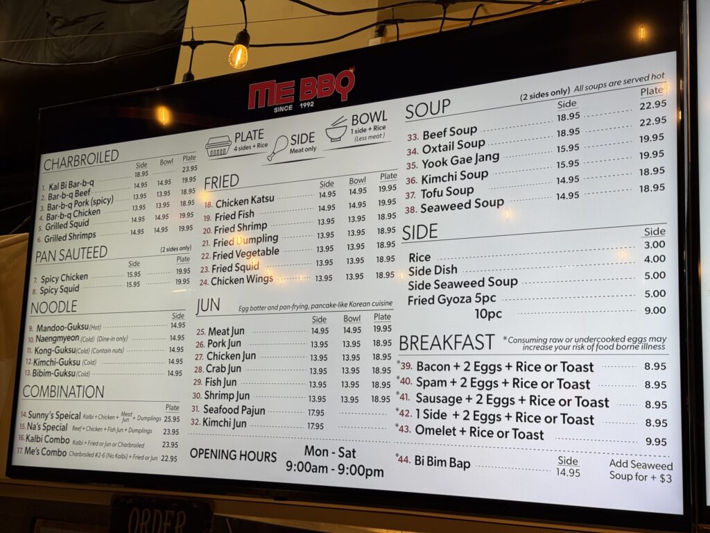 韓国焼肉店「ME BBQ」の電光メニューボード。カルビ、チキン、スープ、麺類など、様々なメニューと価格が英語で表示されている。