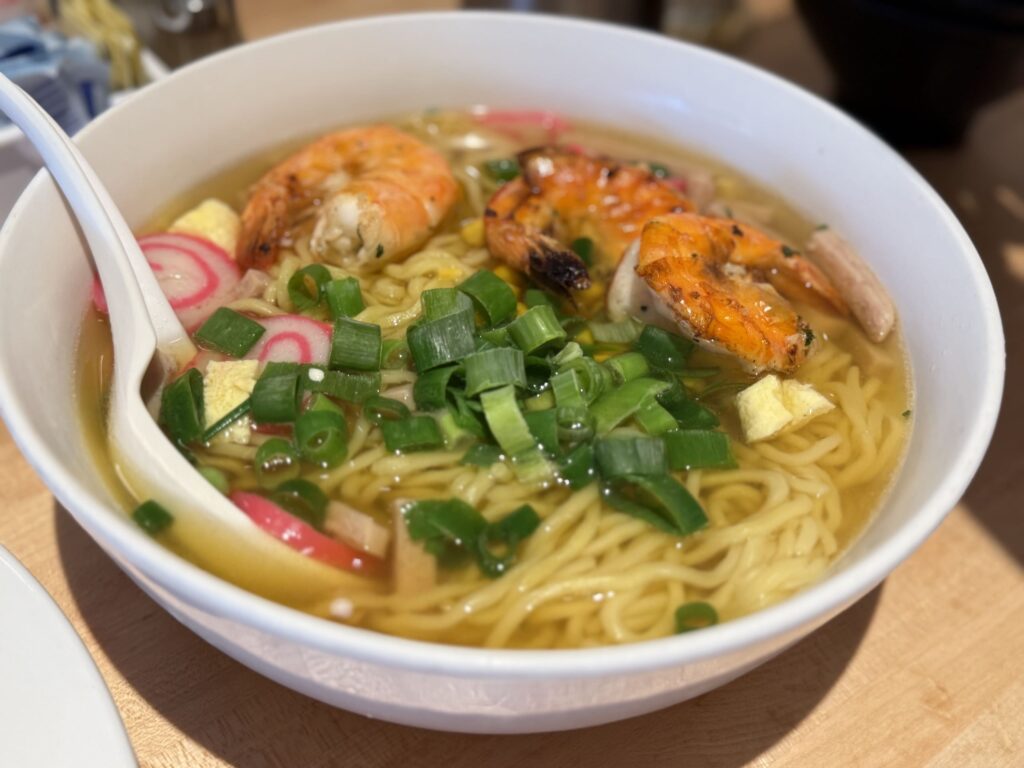 A white bowl of saimin noodle soup topped with grilled shrimp, chopped green onions, narutomaki fish cakes, and egg.