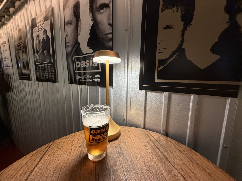 木目調のテーブルの上に置かれた、「oasis live '25」のロゴが入ったビールグラス。背景の波板の壁には、ロックバンドOasisのポスターが複数飾られている。