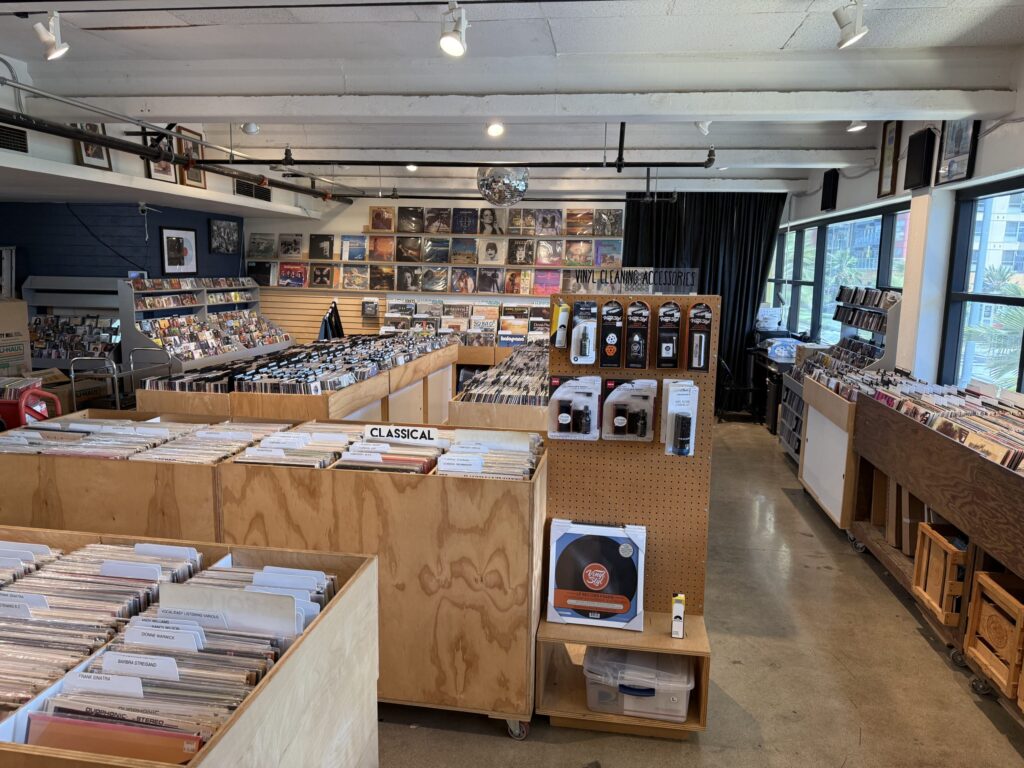 The interior of a spacious record store. Vinyl records are organized by genre in wooden bins, and more records are displayed on the walls. Spotlights and a disco ball are visible on the ceiling.