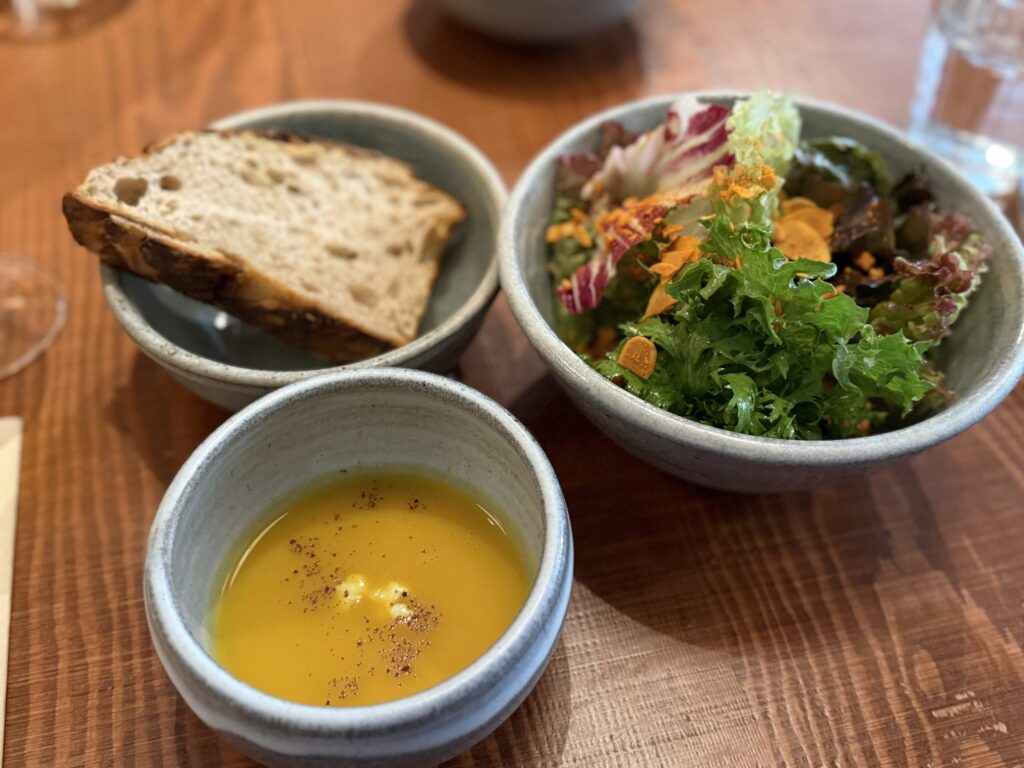 A butternut squash soup, a green salad, and a slice of bread served in rustic ceramic bowls on a wooden table.