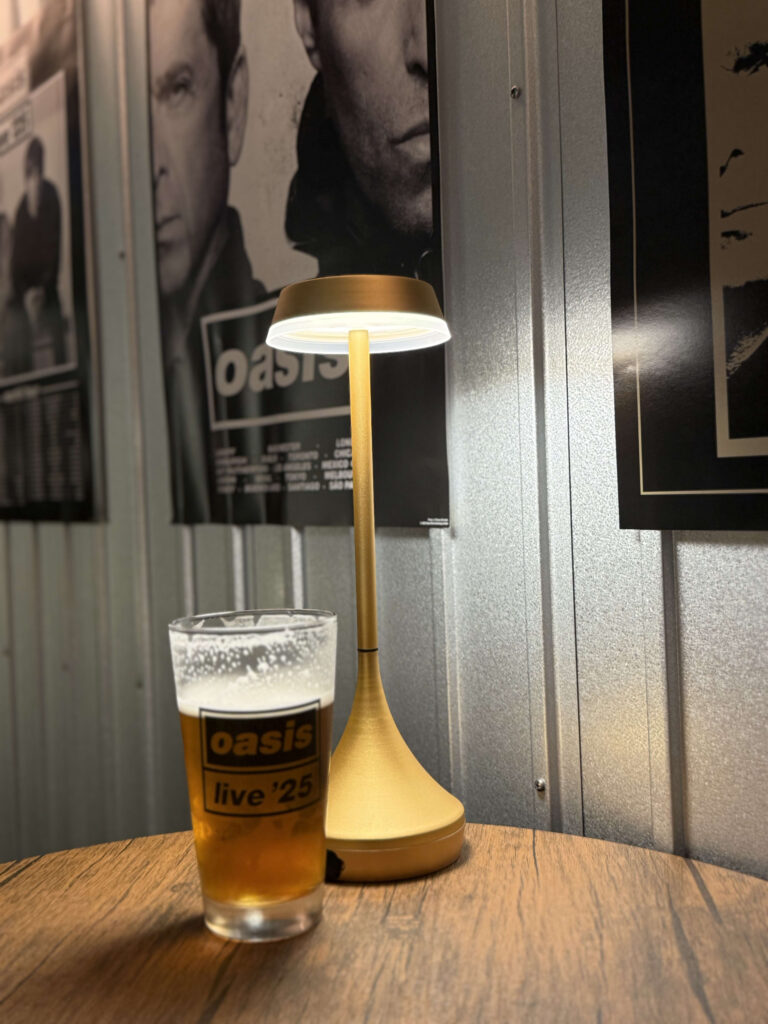 木製のテーブルの上に、オアシスのロゴが入ったビールグラスと金色のテーブルランプが置かれている。背景には波板の壁にオアシスのモノクロポスターが飾られている。