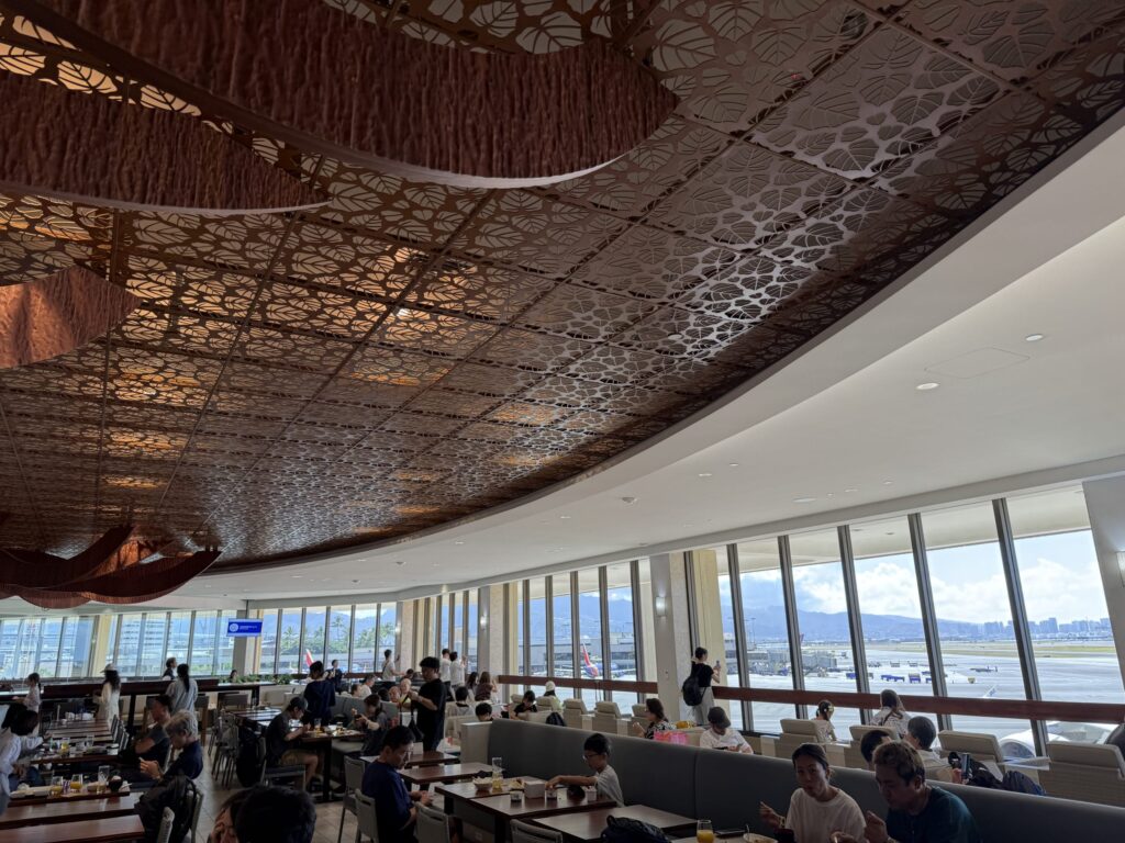 The interior of a spacious airport lounge. Under a ceiling with a leaf pattern design, many people are relaxing, eating, and looking out the window at the airport tarmac.