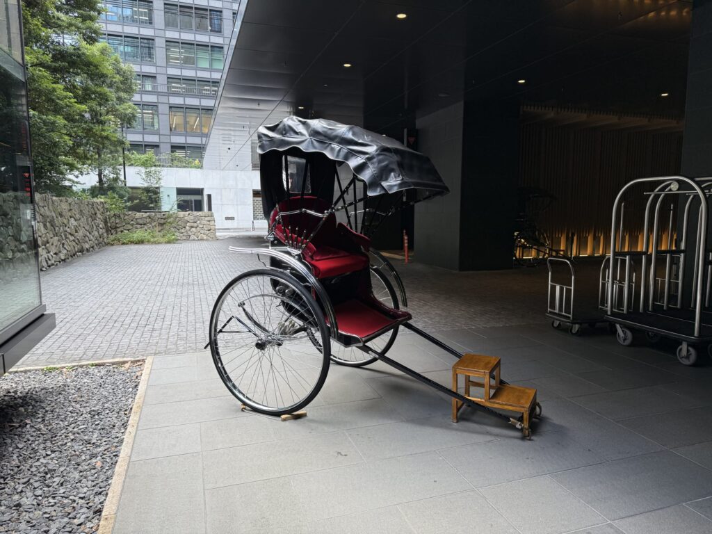 近代的なビルのエントランス前に停められた人力車。黒い幌と赤いベルベットの座席が特徴で、背景にはオフィスビルが見える。
