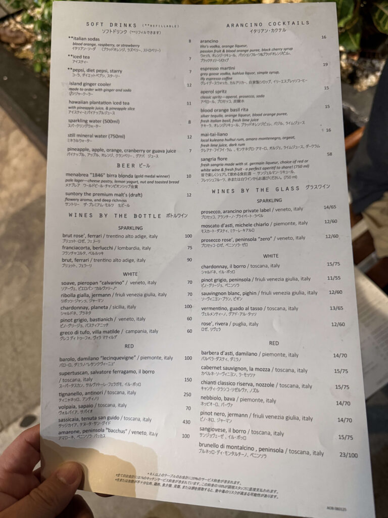 A hand holding the drink menu from the Italian restaurant Arancino. The menu lists soft drinks, cocktails, beer, and wines in both English and Japanese.