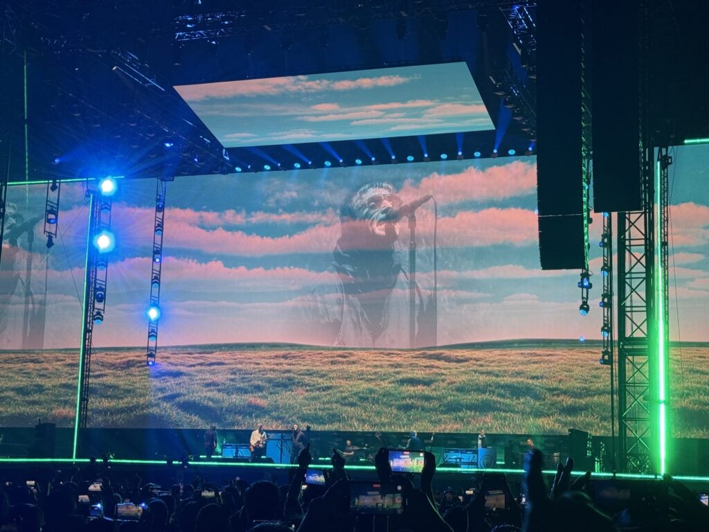 The stage at a Liam Gallagher live concert. A giant screen shows a large image of Liam singing into a microphone, superimposed on a background of a grassy field and a sky with pink clouds. Below the stage, many audience members are holding up their smartphones to record.