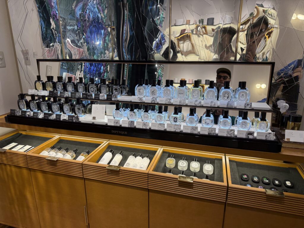 A display shelf in a Diptyque luxury fragrance store. Two tiers of perfume bottles are illuminated, with other products visible in the wooden counter below. A large, fragmented mirror is in the background.