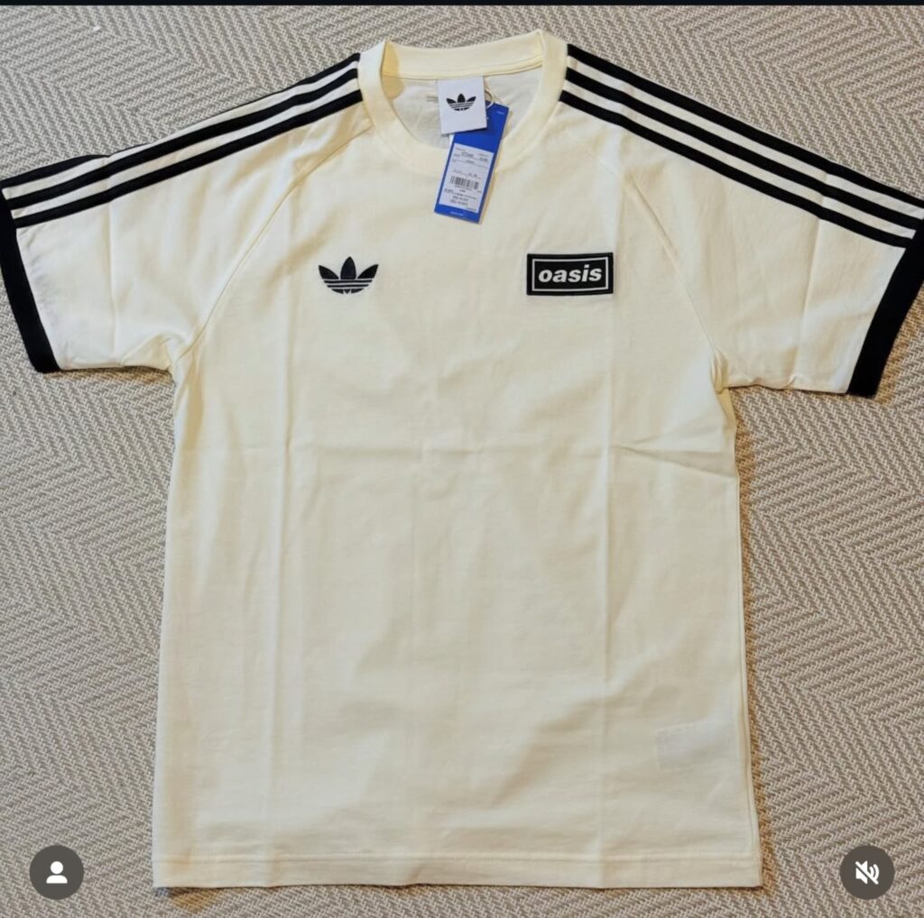 ヘリンボーン柄の床に置かれた、アディダスとオアシスのクリーム色のコラボレーションTシャツ。胸にはアディダスのトレフォイルロゴとオアシスのボックスロゴがあり、肩には黒いスリーストライプスが入っている。