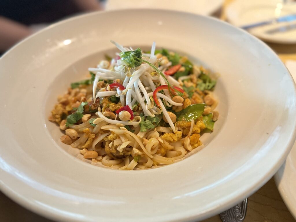 A plate of Pad Thai in a white bowl, topped with bean sprouts, peanuts, and sliced red chili.