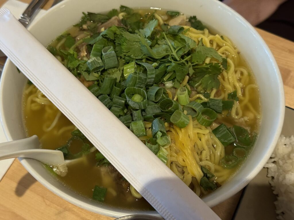 A large white bowl of oxtail soup with ramen noodles, generously topped with chopped scallions and cilantro, with chopsticks in a paper sleeve laid across the bowl.