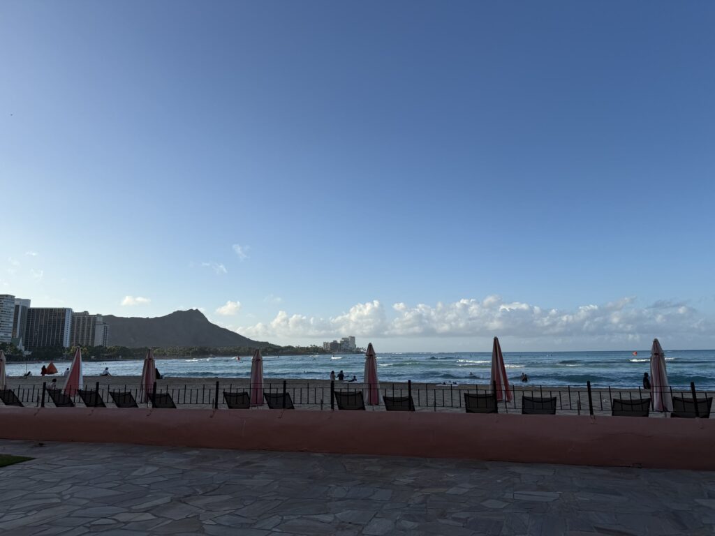 A wide view of Waikiki Beach with Diamond Head in the distance under a clear blue sky. Closed beach umbrellas are lined up on a patio in the foreground.