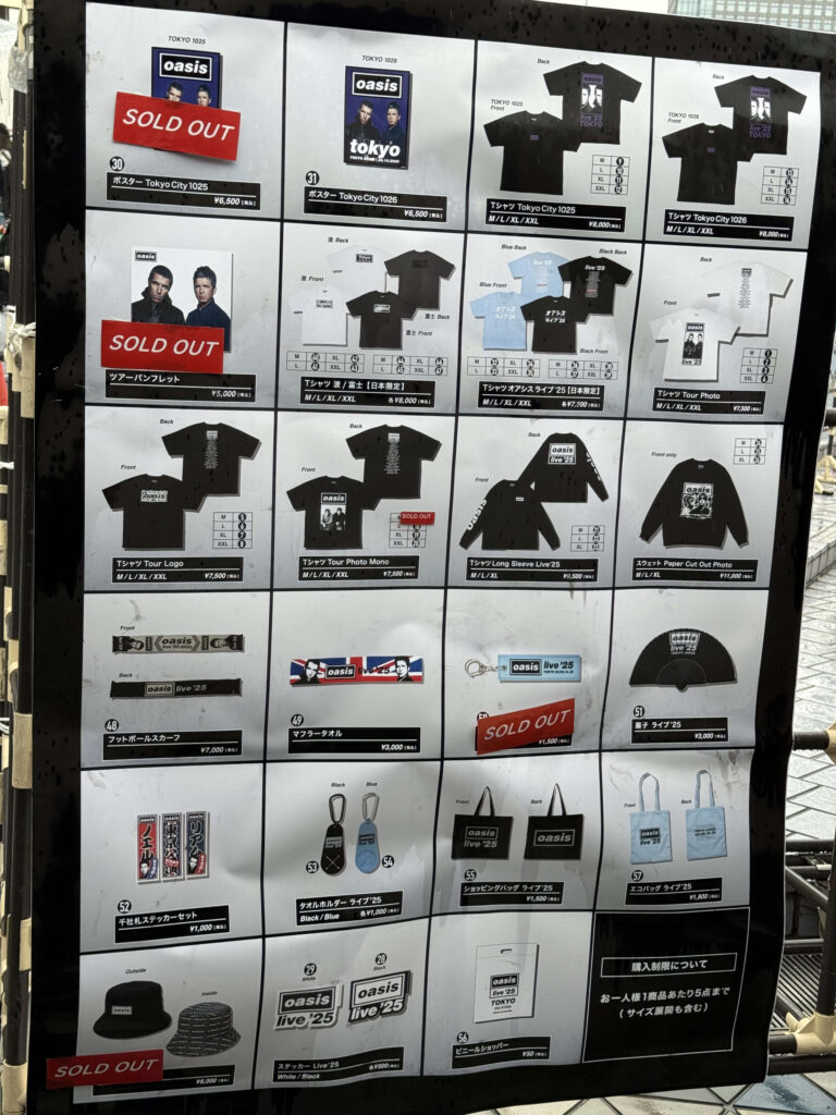 「オアシス ライブ '25」東京公演の物販ブースに掲示されたグッズ一覧ポスター。Tシャツやタオル、ポスターなどの商品写真と価格が掲載されており、一部の商品には「SOLD OUT」の札が貼られている。
