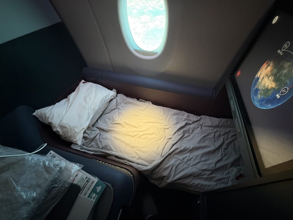 飛行機のビジネスクラスの座席がフルフラットのベッドになっており、枕と掛け布団が用意されている。窓の外には青空と雲が見え、モニターにはフライトマップが表示されている。