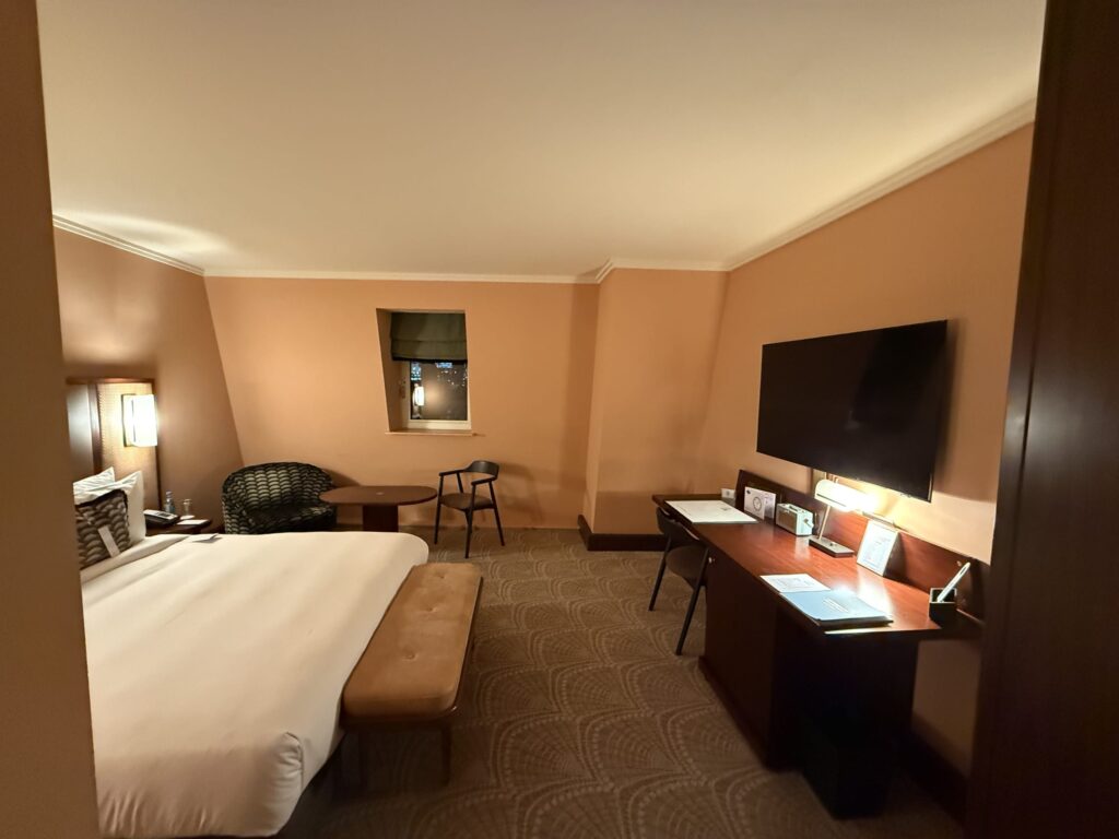 A hotel room at night illuminated by warm lighting. There is a bed on the left, a desk and TV on the right, and a small window and chairs in the back.