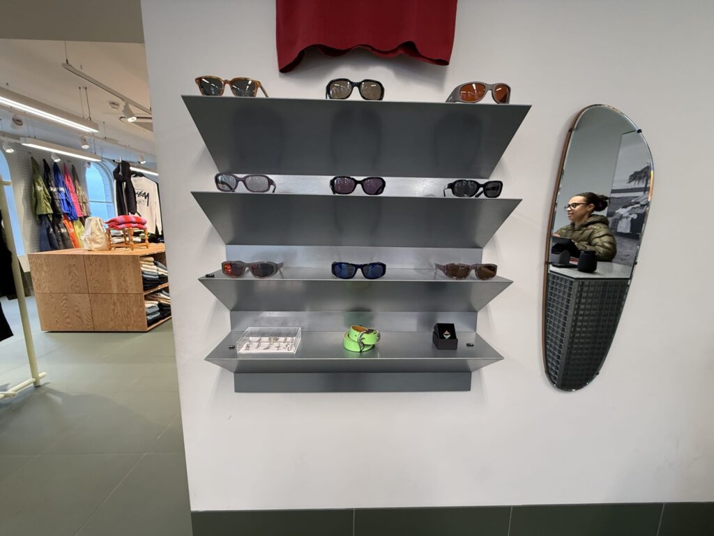 Inside a boutique, various sunglasses are displayed on a modern gray shelf mounted on a white wall. To the right, an oval mirror reflects a woman.