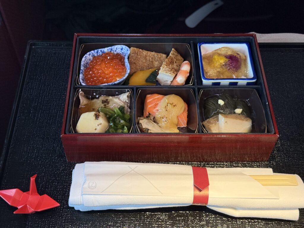 黒いお盆の上に置かれた、6つに仕切られた和食の弁当箱。中にはいくら、焼き魚、煮物など彩り豊かな料理が並んでいる。横には赤い折り鶴と箸が添えられている。