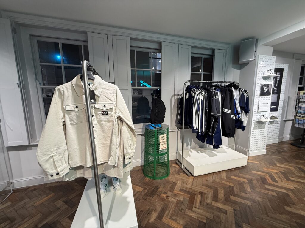 The interior of a clothing store. A white corduroy jacket is displayed in the foreground, with a rack of blue and white tracksuits visible in the background. The floor is a herringbone wood pattern.