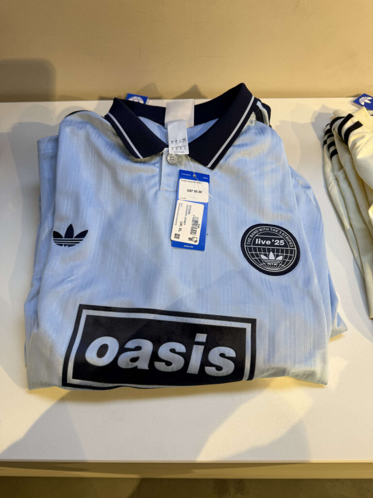 A light blue Adidas x Oasis collaboration polo shirt folded on a shelf. It features the large Oasis logo on the chest and a circular emblem with the Adidas trefoil logo.