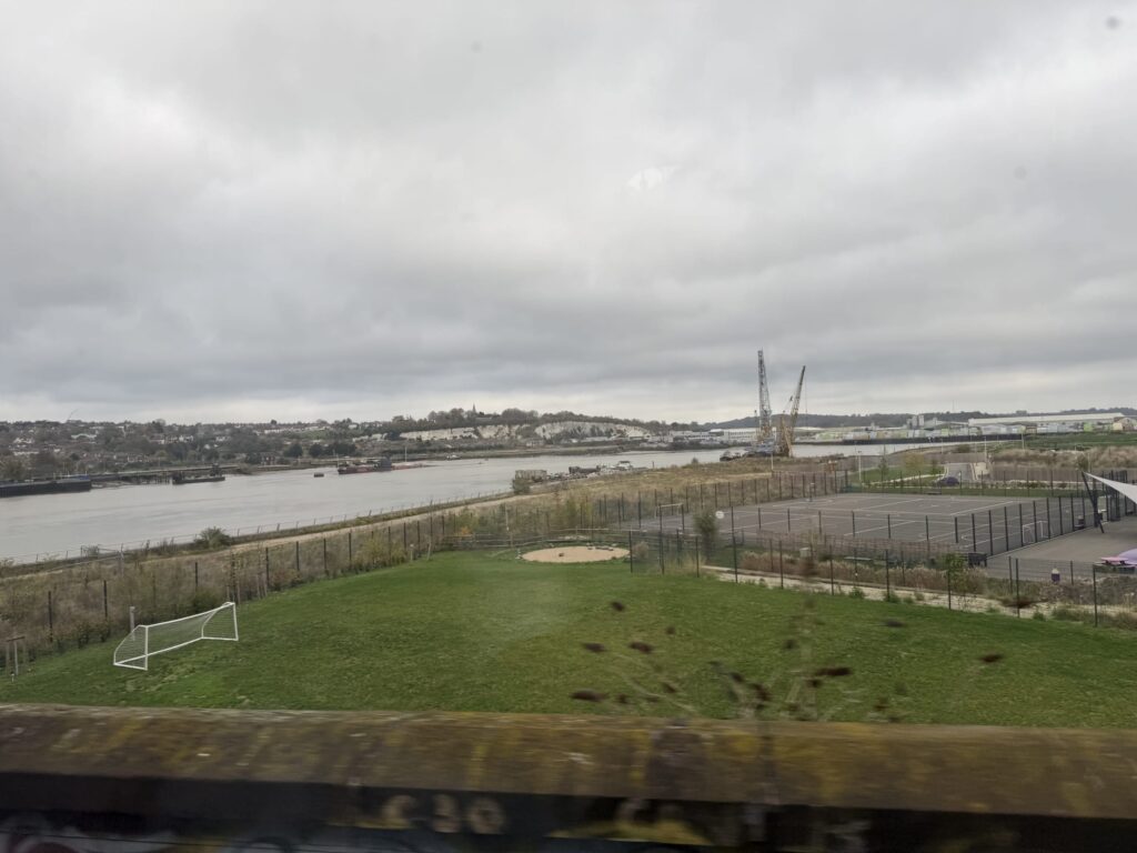 電車の車窓から見た川沿いの風景。曇り空の下、手前にサッカーゴールのある緑地、川の向こう岸にクレーンや町並みが見える。