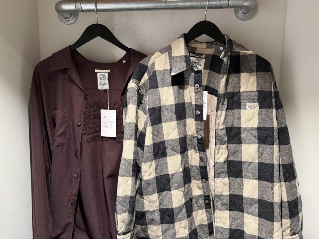 Two shirts hanging on an industrial clothing rack. On the left is a solid maroon shirt, and on the right is a quilted shirt with a black and cream block check pattern.