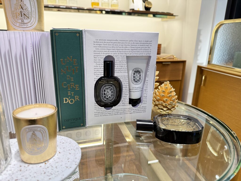 A Diptyque Orphéon gift set displayed on a glass table. The open box reveals an eau de parfum and a hand & body wash, surrounded by a gold candle and a gold pinecone decoration.