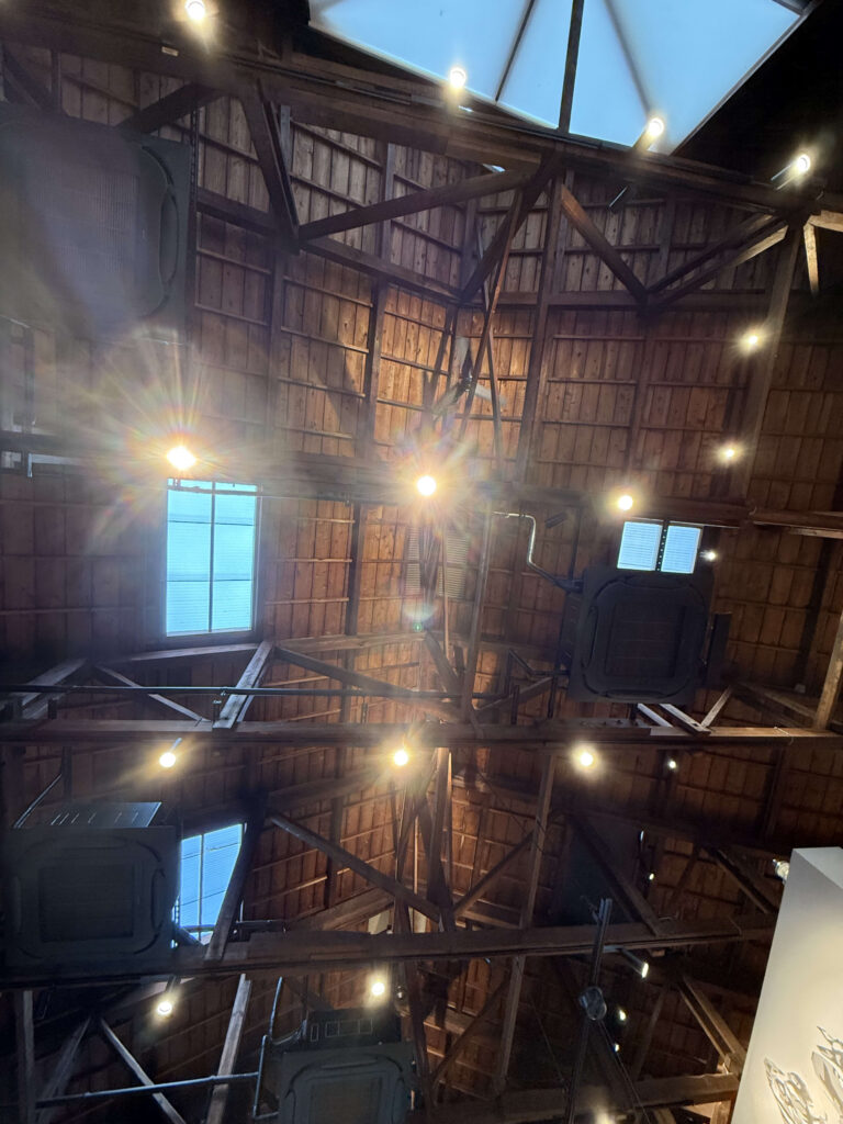 A view looking up at a high, rustic wooden ceiling with exposed beams, trusses, skylights, and warm, glowing lights.