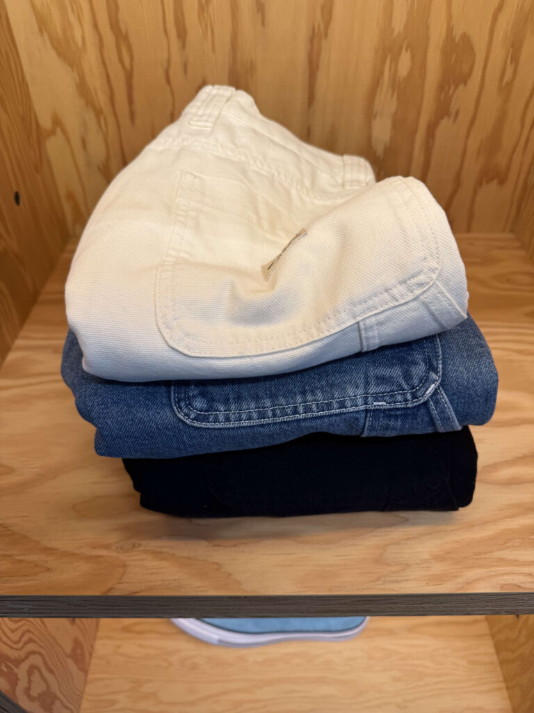 木製の棚の上に、オフホワイト、デニム、ブラックの3色が積み重ねて置かれているStussyのダブルニーパンツ。