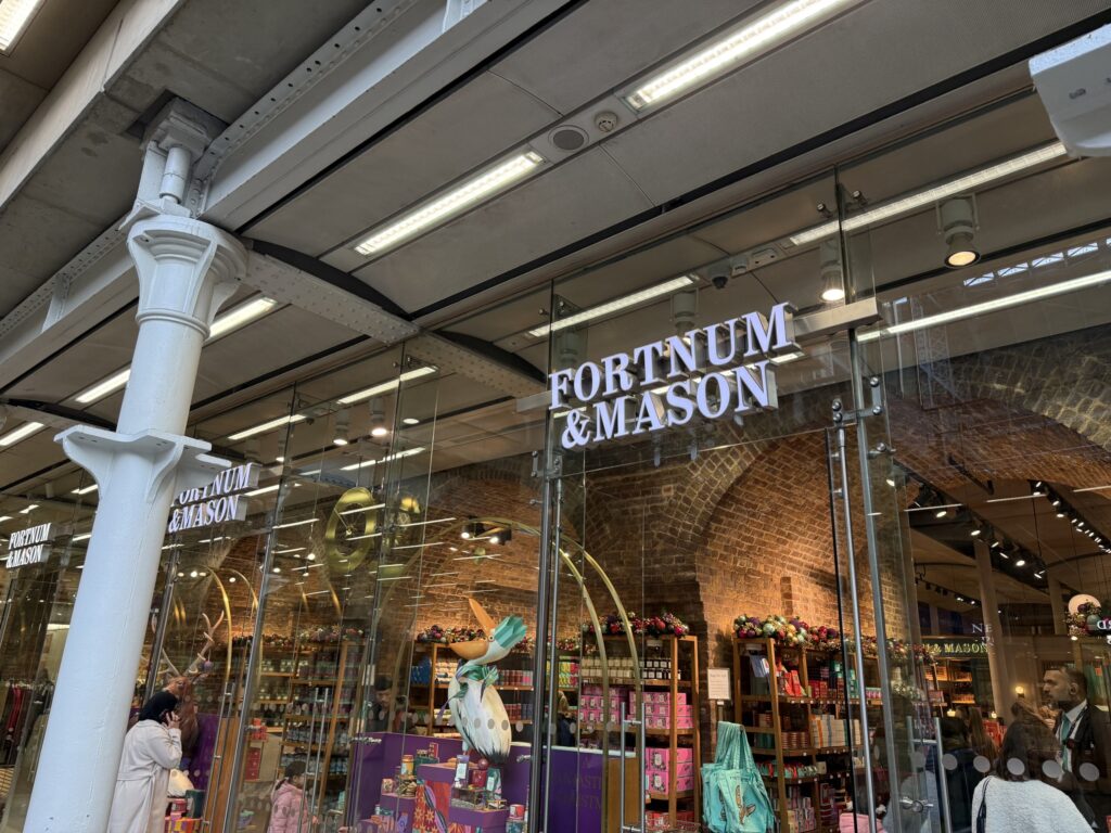 The exterior of a Fortnum & Mason store. A white logo is displayed above the glass facade, and brick walls and product shelves are visible inside the store.