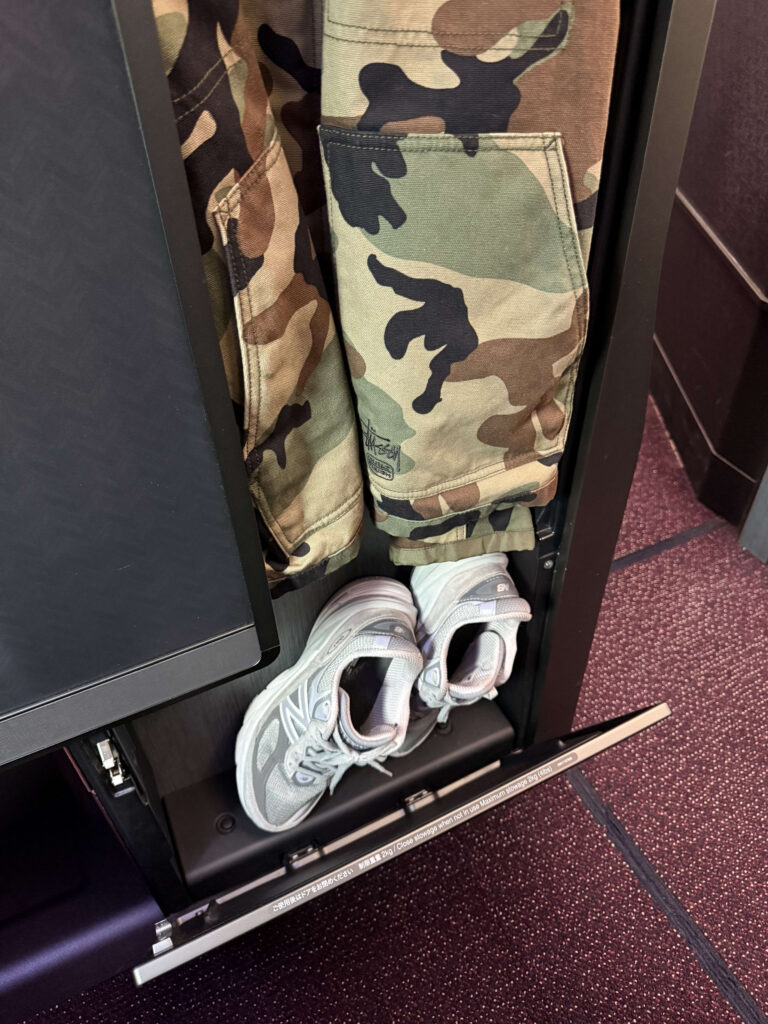 飛行機のビジネスクラスの座席にある収納スペース。迷彩柄のジャケットが掛けられ、足元にはグレーのスニーカーが置かれている。