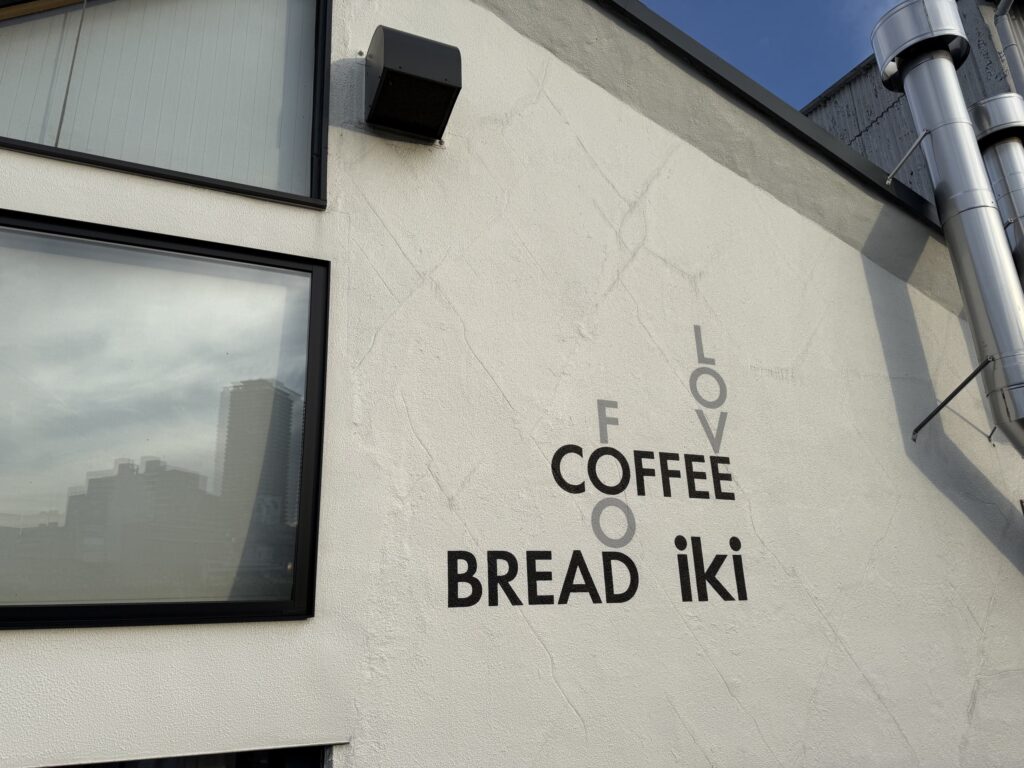 白い壁に「LOVE FOR COFFEE BREAD iki」のロゴがデザインされたカフェ「iki」の外観。左側には黒い窓枠、右上には銀色のダクトが見える。