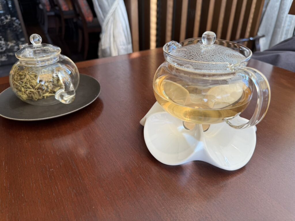 Two glass teapots on a wooden table. The teapot in the foreground contains brewed jasmine tea and is kept warm on a white warmer, while the pot in the background contains tea leaves.