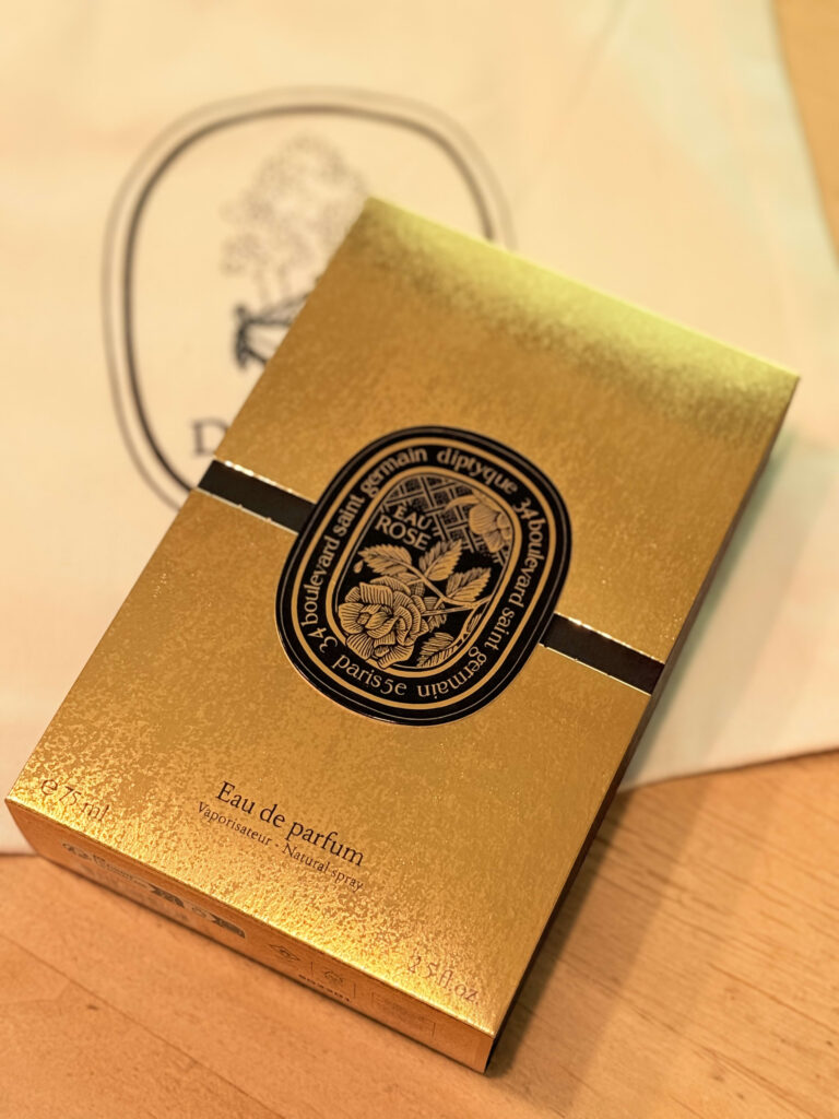 木製のテーブルの上に置かれた、diptyqueの香水「オーローズ オードパルファン」の金色の箱。背景にはブランドのトートバッグが写っている。