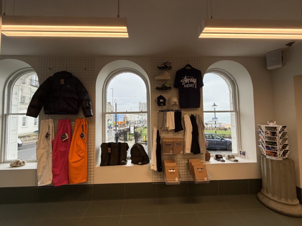 The interior of a clothing store. Merchandise such as jackets, pants, hats, and sneakers are displayed on a wire mesh on a wall with arched windows. A seaside townscape is visible outside the windows.