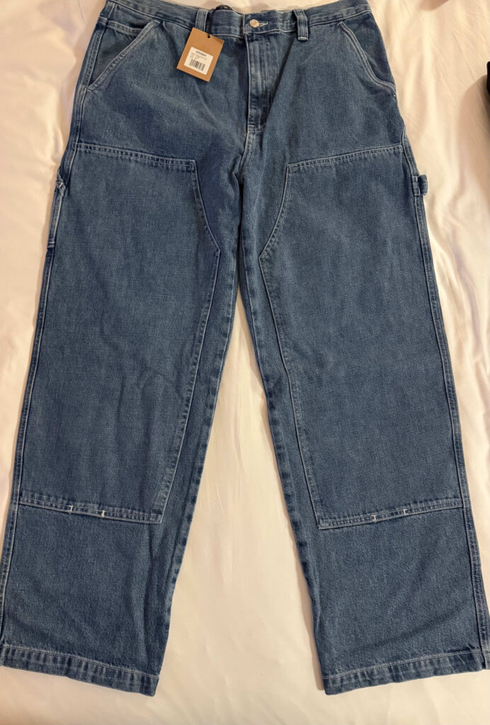 A pair of Stüssy denim painter pants laid flat on a white surface. A brown tag is attached to the waist, and the knees are reinforced with a double-knee design.