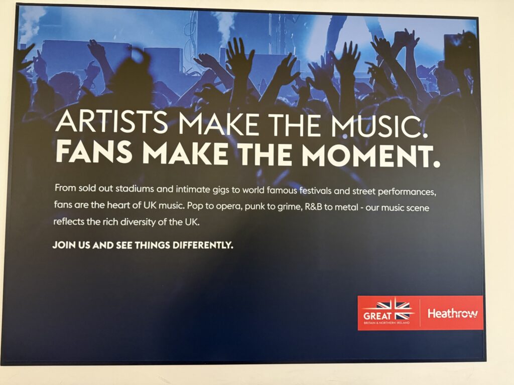 ヒースロー空港に掲示された、イギリスの音楽シーンを宣伝するポスター。コンサートで盛り上がる観客のシルエットを背景に、「ARTISTS MAKE THE MUSIC. FANS MAKE THE MOMENT.(アーティストは音楽を創り、ファンは瞬間を創る)」という白い文字が書かれている。
