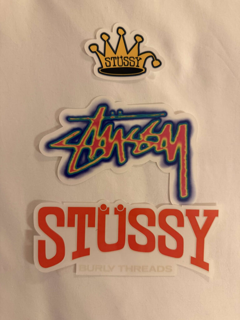 Three different Stüssy stickers on a white background. From the top: a crown logo, a gradient-colored graffiti logo, and a red arched logo.