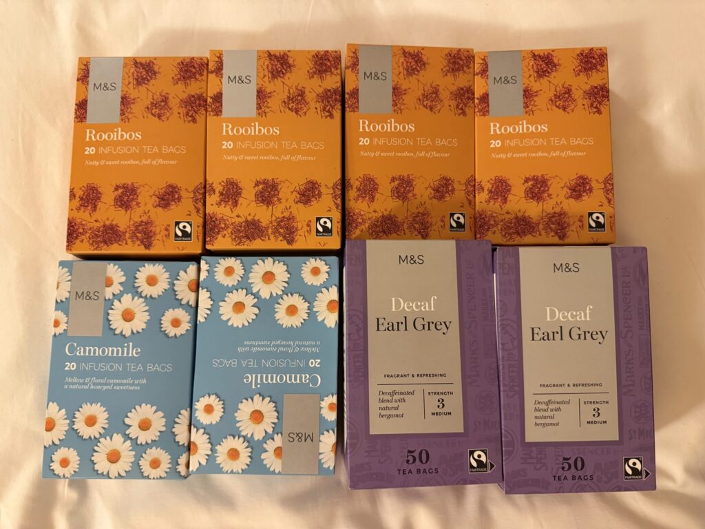 Eight boxes of Marks & Spencer tea bags arranged in two rows. The top row has four orange Rooibos boxes. The bottom row has two blue Camomile boxes and two purple Decaf Earl Grey boxes.