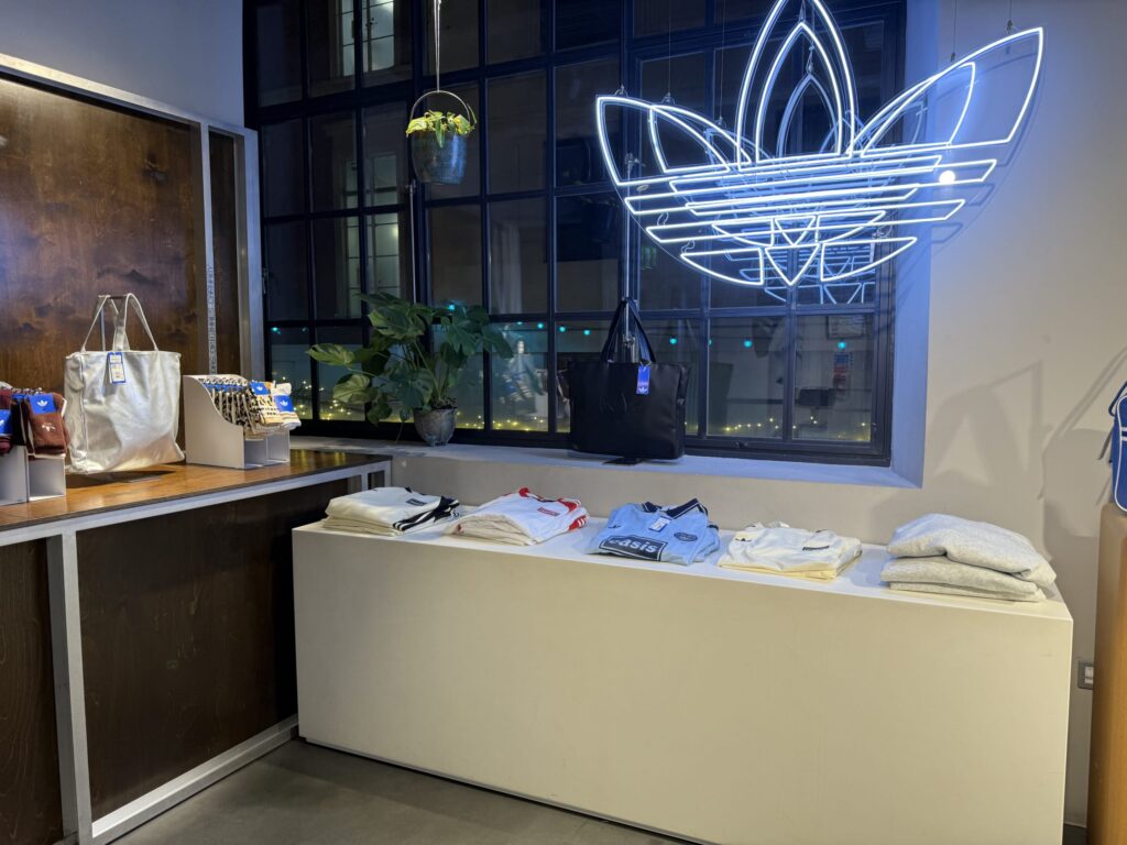 The interior of an Adidas Originals store. A blue neon trefoil logo is displayed by the window, and products like an Oasis collaboration T-shirt are arranged on a white platform.