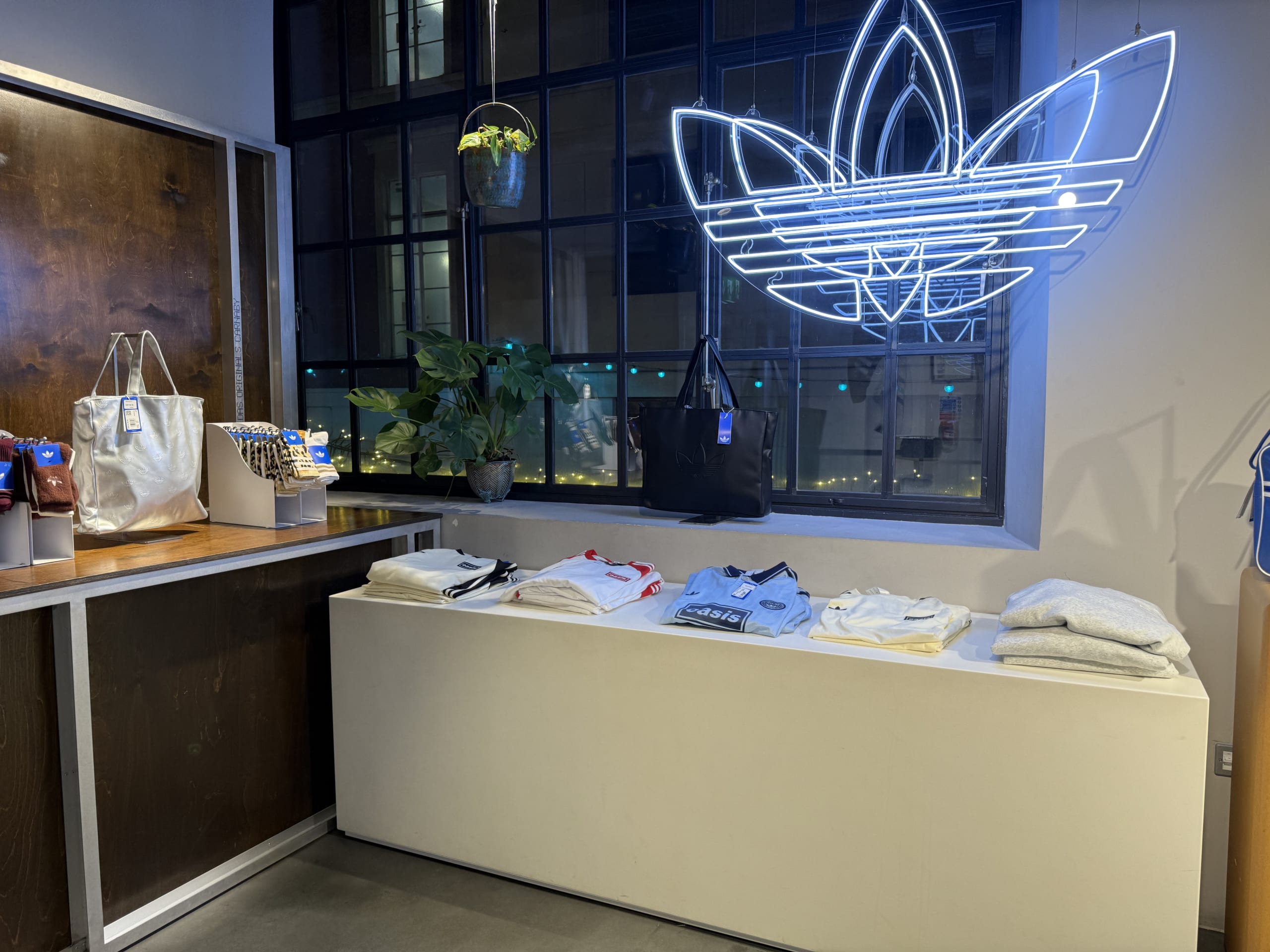 The interior of an Adidas Originals store. A blue neon trefoil logo is displayed by the window, and products like an Oasis collaboration T-shirt are arranged on a white platform.
