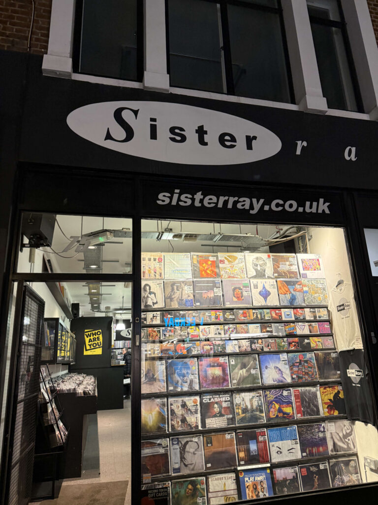 The exterior of the Sister Ray record store at night. Its bright show window is filled with numerous record sleeves covering the entire wall.
